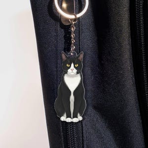 Puede incluir: Un llavero de gato blanco y negro con un llavero plateado. El gato está sentado con los ojos amarillos y el pecho blanco. El llavero está sujeto a una bolsa negra.