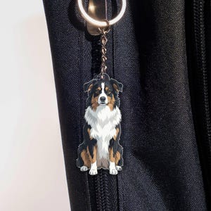 May include: A keychain featuring a sitting Australian Shepherd dog. The dog has black, brown, and white markings. The keychain is attached to a silver-colored ring and a black cord. The background is a black fabric.
