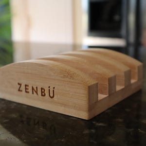 May include: A light brown bamboo soap dish with the engraved word "ZENBÜ". The dish has a curved top with five slots for soap bars. The design is simple and modern, suitable for a bathroom or kitchen.