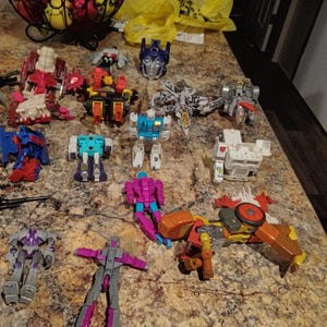 May include: A collection of colorful Transformers action figures, including robots in various colors such as red, blue, white, purple, and orange. The toys are arranged on a speckled countertop, showcasing their detailed designs and transforming capabilities.