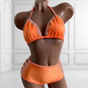May include: A bright orange two-piece bikini set featuring a halter-style top and high-waisted bottoms. The fabric is adorned with scattered iridescent crystals and intricate crystal-lined trim along the edges and side cutouts of the bottoms.