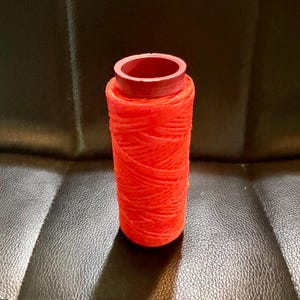 May include: A spool of bright red thread, tightly wound around a cylindrical core. The thread is a vibrant, solid color, and the spool has a matching red top. The image is a close-up shot, highlighting the texture and color of the thread.