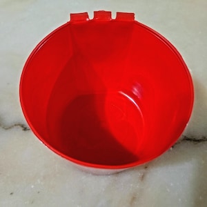 Może przedstawiać: Żywy czerwony plastikowy pojemnik o zaokrąglonym kształcie i trzech małych wypustkach na krawędzi. Pojemnik ma gładkie, błyszczące wykończenie i wydaje się pusty. Pojemnik stoi na białej i szarej powierzchni.