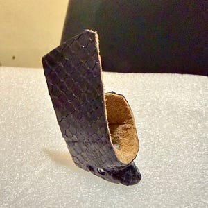 Puede incluir: Una funda de cuero morado oscuro con un patr&oacute;n de escamas. La funda tiene una forma curva con un interior marr&oacute;n claro y tres peque&ntilde;os agujeros cerca de la parte inferior. El producto es probablemente para llevar un objeto peque&ntilde;o.