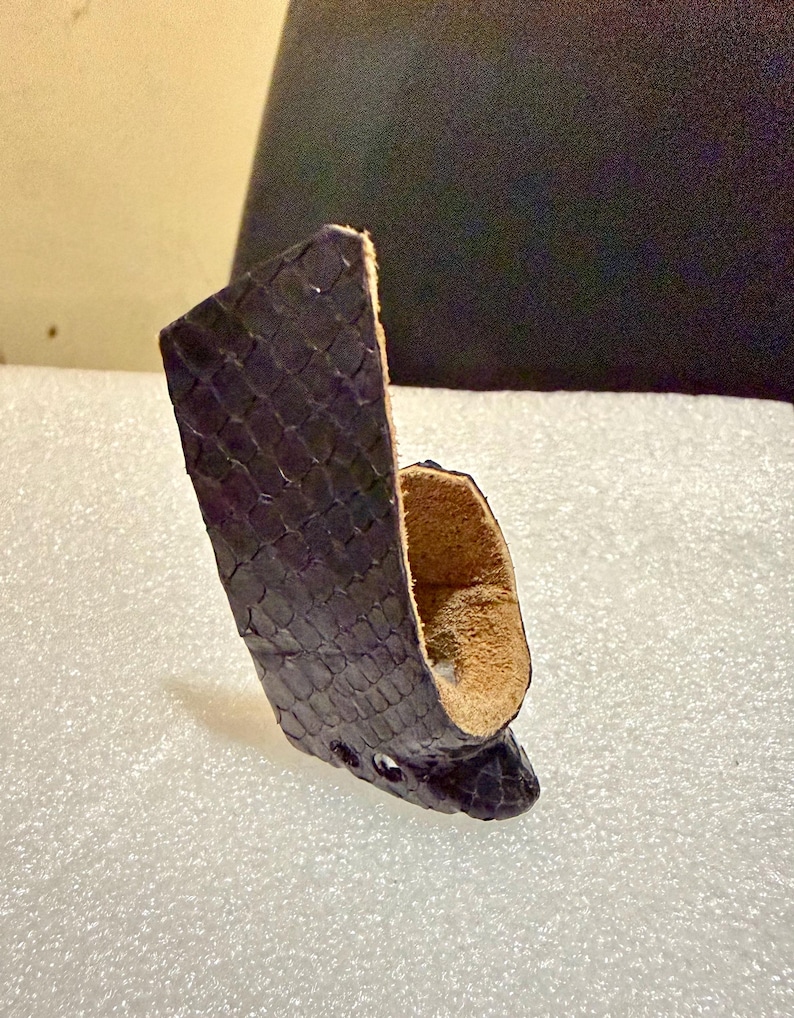 Puede incluir: Una funda de cuero morado oscuro con un patr&oacute;n de escamas. La funda tiene una forma curva con un interior marr&oacute;n claro y tres peque&ntilde;os agujeros cerca de la parte inferior. El producto es probablemente para llevar un objeto peque&ntilde;o.