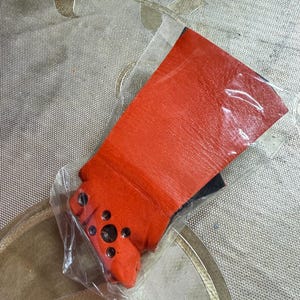 Puede incluir: Un art&iacute;culo de cuero naranja brillante, posiblemente un guante o parte de una prenda, est&aacute; envuelto en pl&aacute;stico transparente. El cuero tiene una superficie texturizada y es de un tono rojo-naranja vibrante. El art&iacute;culo tiene varios peque&ntilde;os detalles circulares oscuros.