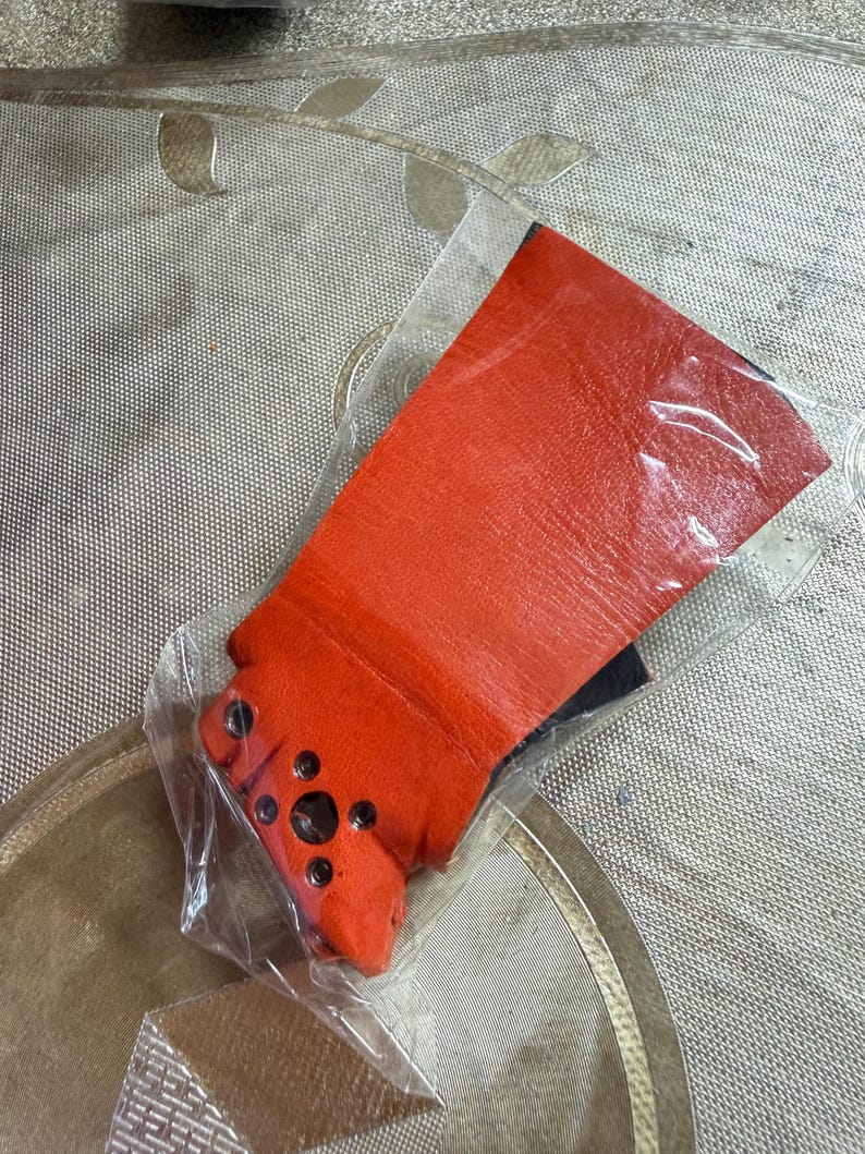 Puede incluir: Un art&iacute;culo de cuero naranja brillante, posiblemente un guante o parte de una prenda, est&aacute; envuelto en pl&aacute;stico transparente. El cuero tiene una superficie texturizada y es de un tono rojo-naranja vibrante. El art&iacute;culo tiene varios peque&ntilde;os detalles circulares oscuros.