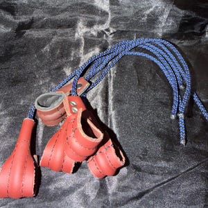 May include: Red leather hand restraints with black stitching and a blue and black rope. The restraints have a finger loop design and are connected to a leather wrist cuff. The background is a dark, textured fabric.