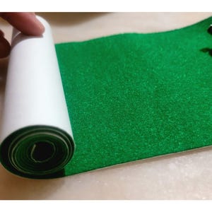 May include: A roll of bright green felt material with a white backing is partially unrolled, revealing the felt's texture. The felt is a vibrant, solid green color. The image shows a close-up of the felt roll.