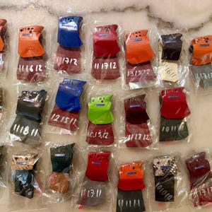 Puede incluir: Una colecci&oacute;n de peque&ntilde;os accesorios de cuero de colores, cada uno empaquetado individualmente en pl&aacute;stico transparente. Los art&iacute;culos presentan varios colores, incluyendo naranja, azul, rojo y marr&oacute;n, con algunos mostrando patrones texturizados. Cada paquete tiene n&uacute;meros escritos a mano.