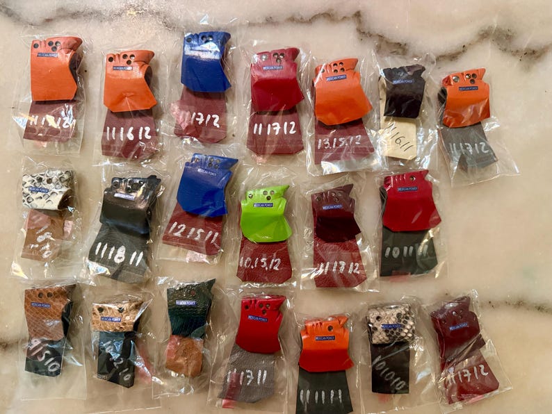 Puede incluir: Una colecci&oacute;n de peque&ntilde;os accesorios de cuero de colores, cada uno empaquetado individualmente en pl&aacute;stico transparente. Los art&iacute;culos presentan varios colores, incluyendo naranja, azul, rojo y marr&oacute;n, con algunos mostrando patrones texturizados. Cada paquete tiene n&uacute;meros escritos a mano.