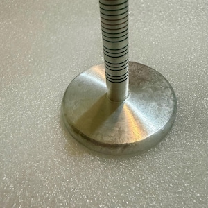 May include: A silver-colored metal bottle stopper with a round base and a cylindrical handle. The handle has thin, dark green stripes. The stopper is designed to seal bottles.
