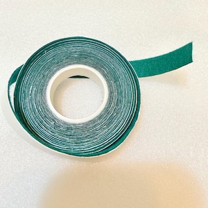 May include: A roll of green double-sided tape with a white core. The tape is unrolled slightly, revealing the adhesive side. The background is a white, textured surface, and a red roll of tape is visible in the background.