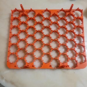 May include: An orange plastic battery holder with multiple circular compartments. The holder has a rectangular shape with raised edges and several small, cylindrical protrusions. The design suggests it's for organizing and storing batteries.