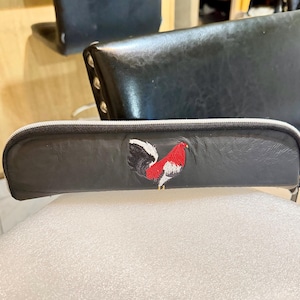 May include: A black pencil case with a white zipper and an embroidered rooster design in red, white, and black. The case is rectangular and appears to be made of leather or a similar material.