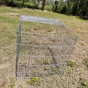 Puede incluir: Una jaula rectangular de alambre metálico con un panel superior abatible y una puerta frontal. La estructura está construida con barras de acero de color plateado dispuestas en forma de rejilla, creando un recinto abierto con pestillos.