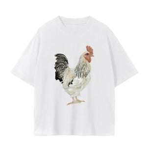 May include: White t-shirt featuring a detailed watercolor illustration of a rooster. The rooster has a red comb and wattle, with black and white feathers. The shirt is a casual, everyday wear item.