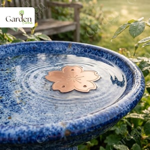 May include: A blue ceramic bird bath with a copper flower-shaped floating ornament. The bird bath has a speckled design and is filled with water. The background includes greenery and a wooden bench.