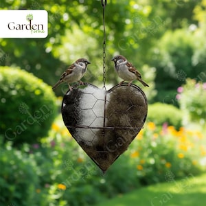 May include: A heart-shaped bird feeder with a wire mesh design, divided into two sections, one white and one brown. Two small birds perch on top. The feeder hangs from a chain against a blurred green background.