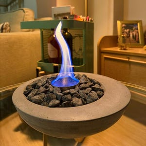 Peut inclure: Un brasero en béton rempli de pierres gris foncé, avec une flamme bleue et blanche brûlant au centre. Le brasero est posé sur une surface en verre, avec un arrière-plan flou d'une pièce.
