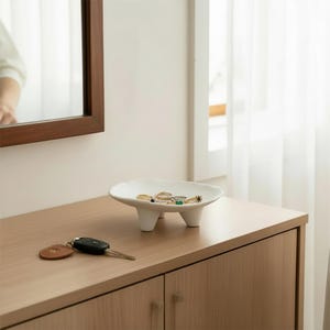 May include: A white ceramic jewelry dish with three small legs, holding various rings. The dish sits on a light wood surface, next to a set of keys. A brown-framed mirror is visible in the background.