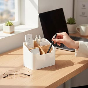 Op de afbeelding: Een witte bureau-organizer met houten verdelers, met een afstandsbediening, pennen en andere kantoorbenodigdheden. Een persoon houdt een zwarte pen vast. Een laptop en een bril staan ook op het houten bureau.