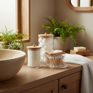 May include: A bathroom set comprising three glass jars with bamboo lids. One jar contains cotton pads, another cotton wool balls, and the third cotton buds. The jars are displayed on a wooden surface, with a neutral backdrop.