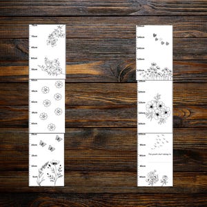 May include: Two white growth charts with black floral and butterfly illustrations, set against a dark wood background. Each chart is marked with centimeter increments. One chart reads up to 70cm, the other up to 150cm. The text "The growth chart belongs to" is visible.