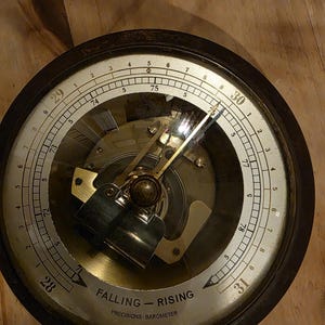 May include: A vintage barometer with a dark wooden frame and a clear glass face. The dial features numbers and markings for pressure readings, with the words "FALLING - RISING" and "PRECISIONS-BAROMETER" visible. The barometer is marked "WEST GERMANY".