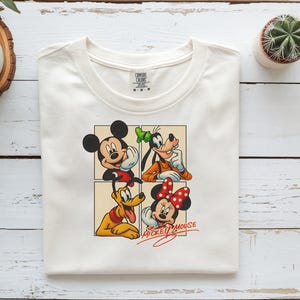 以下が含まれることがあります： クリーム色の半袖Tシャツ。ミッキーマウス、グーフィー、プルート、ミニーマウスが4つのパネルのグリッドに配置されたグラフィックプリントが施されています。前面のキャラクターの下には、赤い筆記体でMickey Mouseの文字がプリントされています。