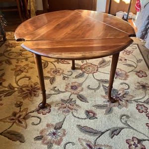 May include: A round, wooden table with a warm, brown finish. The table has a drop-leaf design and four curved legs. The table is set on a floral patterned rug.
