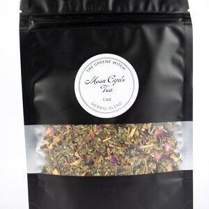 May include: A black pouch of herbal tea with a clear window showing the tea blend. The tea blend includes green herbs and pink rose petals. The pouch has a white circular label with the text "The Greene Witch Moon Cycle Tea 1 oz Herbal Blend."
