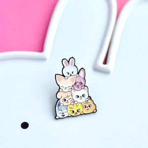 May include: A colorful enamel pin featuring a stack of cartoon animal characters, including a bunny, fox, and chick. The pin has a black outline and is set against a white and pink background. The animals are in various colors.