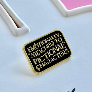May include: A rectangular black enamel pin with gold trim. The pin reads "Emotionally Attached to Fictional Characters" in gold lettering. The pin is set against a white and pink background.