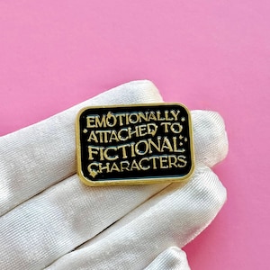 May include: A rectangular black enamel pin with gold trim and the text "EMOTIONALLY ATTACHED TO FICTIONAL CHARACTERS" in gold lettering. The pin is displayed on a white glove against a pink background.