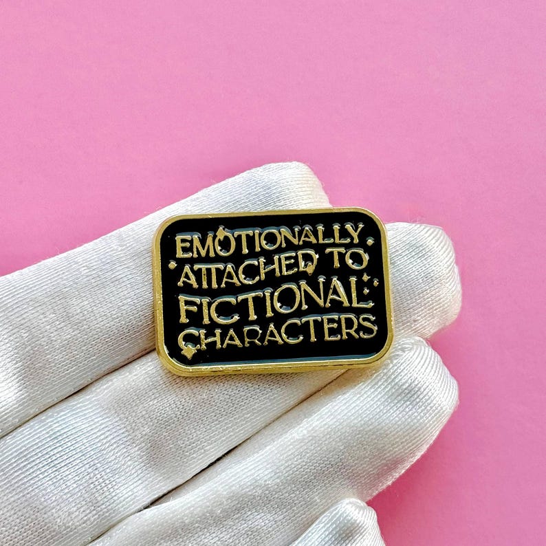 May include: A rectangular black enamel pin with gold trim and the text "EMOTIONALLY ATTACHED TO FICTIONAL CHARACTERS" in gold lettering. The pin is displayed on a white glove against a pink background.