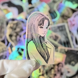 May include: Holographic sticker featuring a manga-style illustration of a woman with long, straight hair and a delicate expression. The sticker has a pearlescent, iridescent finish, with a soft color gradient. The background shows other stickers.