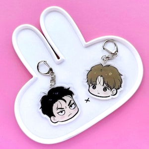May include: Two cartoon character keychains with silver clasps rest on a white bunny-shaped tray against a pink background. One keychain features a character with black hair and an angry expression. The other has a character with brown hair.
