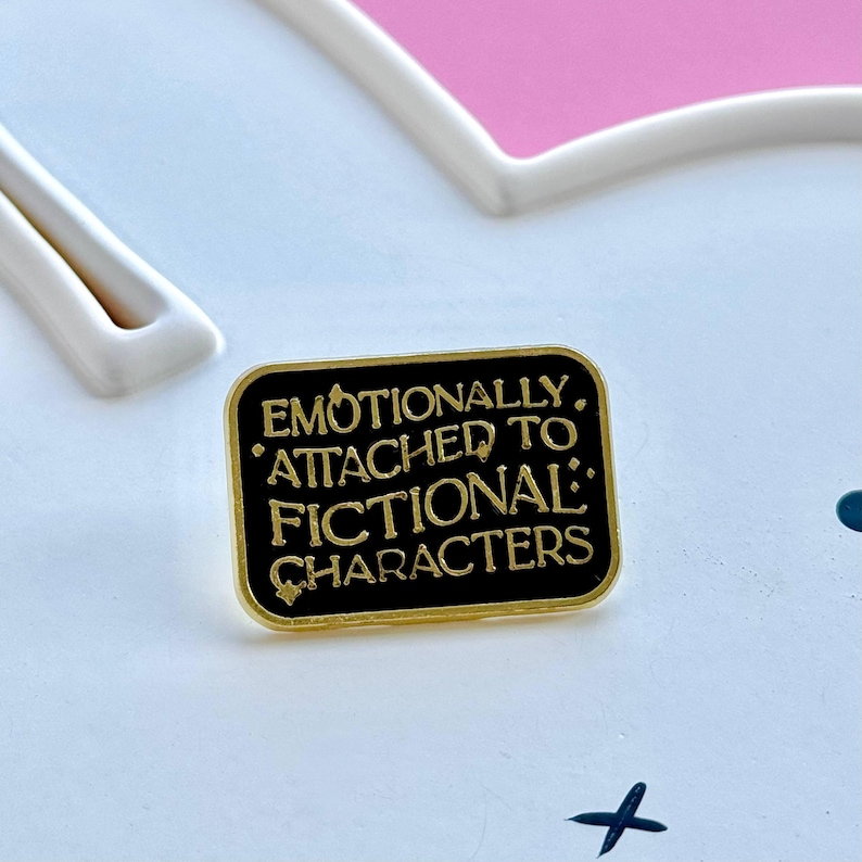 May include: A rectangular black enamel pin with gold trim. The pin features the text "Emotionally Attached to Fictional Characters" in gold lettering. The pin is on a white surface with a pink heart-shaped design.