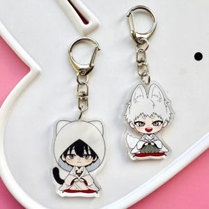 Puede incluir: Dos llaveros de estilo anime con herrajes metálicos plateados. Un llavero presenta un personaje con un atuendo blanco con capucha, cabello negro y una cola negra. El otro llavero representa un personaje con cabello blanco y orejas de zorro, con un atuendo blanco.