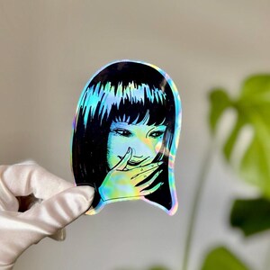 May include: Holographic sticker featuring a manga-style illustration of a woman with black hair and a hand covering her mouth. The sticker has a rainbow iridescent effect, reflecting various colors. The design is a stylized, artistic representation.