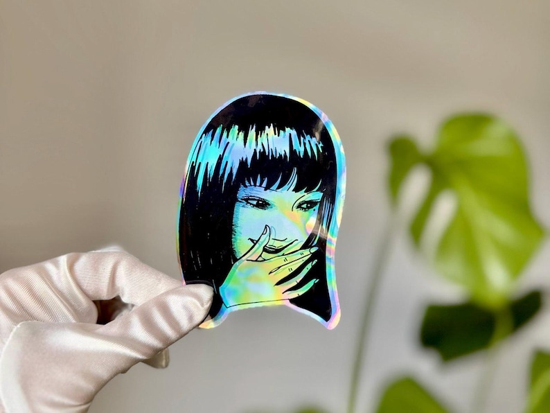 May include: Holographic sticker featuring a manga-style illustration of a woman with black hair and a hand covering her mouth. The sticker has a rainbow iridescent effect, reflecting various colors. The design is a stylized, artistic representation.