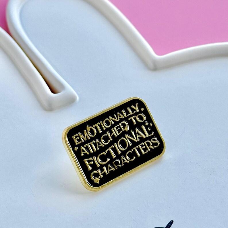 May include: A rectangular black enamel pin with gold trim. The pin reads "Emotionally Attached to Fictional Characters" in gold lettering. The pin is set against a white and pink background.