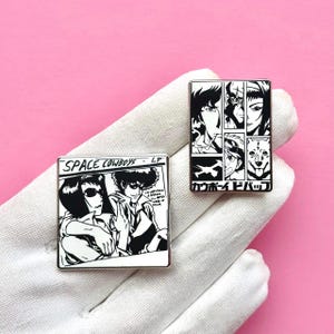 以下が含まれることがあります： 白黒イラストが描かれた2つのエナメルピン。1つのピンには「SPACE COWBOYS LP」と書かれており、2人のキャラクターが描かれています。もう1つのピンには、キャラクターと日本語のテキストが描かれた複数のパネルがあります。どちらのピンも銀色の縁取りがあり、ピンク色の背景にあります。