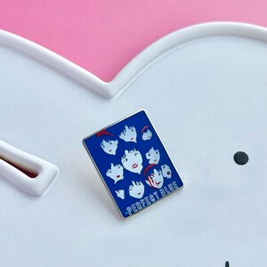 Pode incluir: Um alfinete de esmalte azul com uma borda prateada, apresentando v&aacute;rios rostos estilizados com detalhes em vermelho. O alfinete tem o texto "PERFECT BLUE" em branco. O alfinete est&aacute; sobre um fundo branco e rosa.