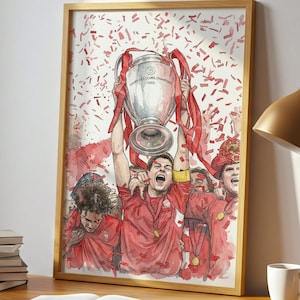 May include: A framed watercolor print of a soccer team celebrating a victory, holding a trophy. The artwork is in a gold frame and sits on a wooden desk with a lamp, books, and a mug. A window and a plant are in the background.