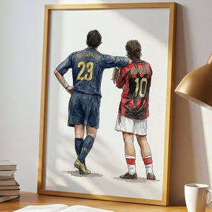 Könnte beinhalten: Ein gerahmter Druck von zwei Fußballspielern in Trikots, einer marineblau mit der Nummer 23, der andere rot und schwarz mit der Nummer 10. Der Druck steht auf einem Holzschreibtisch mit Lampe, Büchern und einer Tasse.