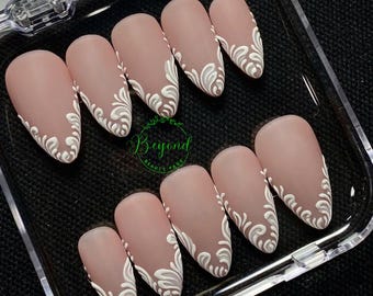 Op de afbeelding: Een set amandelvormige, matte nude press-on nagels met witte bloemmotieven op de punten. De nagels zijn gerangschikt in een doorzichtige plastic doos, met de tekst "Beyond Beauty Page" zichtbaar.