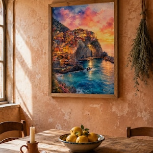 May include: A framed wall art print depicting a coastal village built into a cliffside at sunset, featuring warm orange and pink clouds, illuminated buildings, and deep blue water, displayed in a wooden frame on a textured peach-coloured wall.