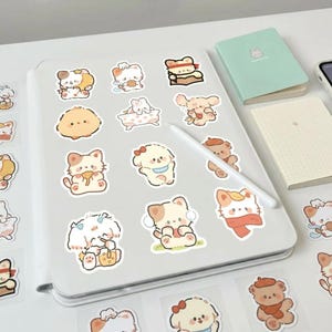 May include: A selection of colourful cartoon stickers depicting adorable animal characters such as cats, dogs, and bears. The stickers are displayed on a white surface alongside a digital tablet, a stylus, and two small notebooks.